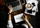 Personne travaillant sur un bureau avec un ordinateur portable, un carnet de croquis, un smartphone et une tasse de café.