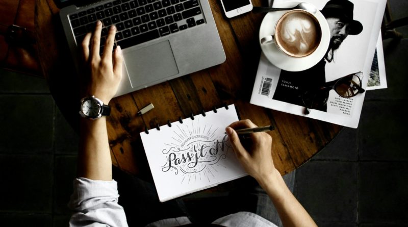 Personne travaillant sur un bureau avec un ordinateur portable, un carnet de croquis, un smartphone et une tasse de café.