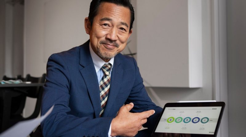 Homme en costume bleu souriant et pointant un graphique circulaire affiché sur une tablette lors d’une présentation professionnelle.