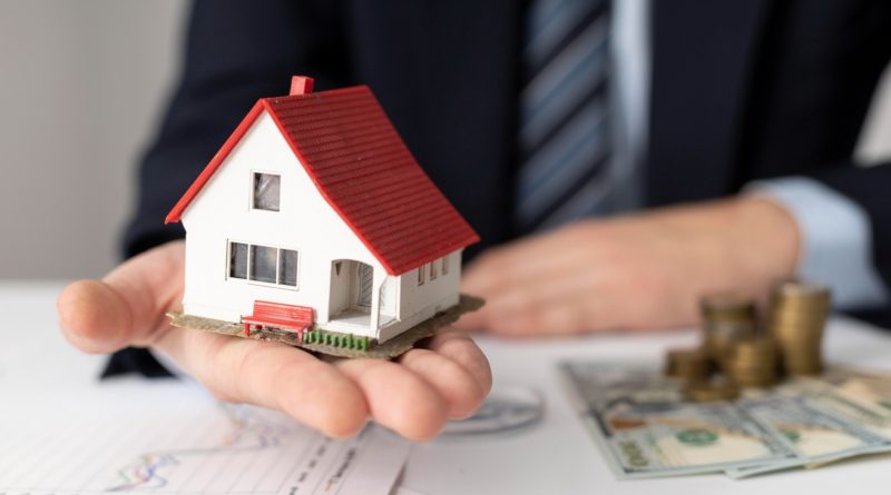 Une main tenant une maison miniature, avec de l’argent et des papiers posés sur une table