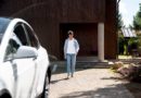Homme marchant devant une maison en bois avec une voiture blanche en charge.