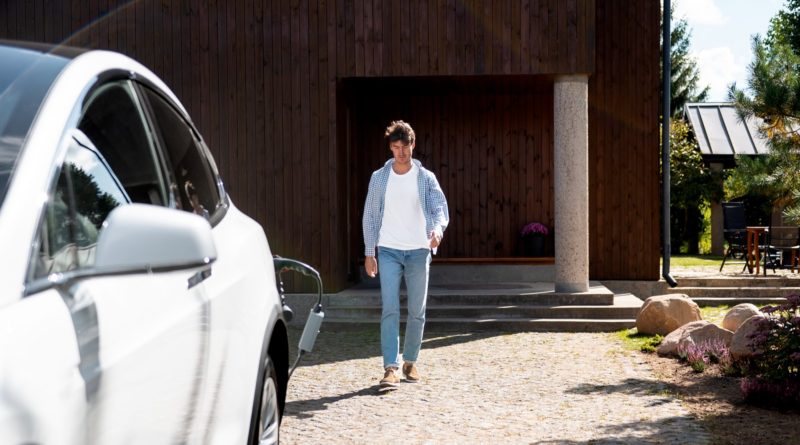 Homme marchant devant une maison en bois avec une voiture blanche en charge.
