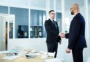 Deux hommes en costume se serrent la main dans un bureau lumineux, avec une table et un ordinateur devant eux