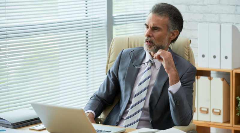 Un homme d'affaires mûr et réfléchi est assis à son bureau devant un ordinateur portable, tenant ses lunettes à la main