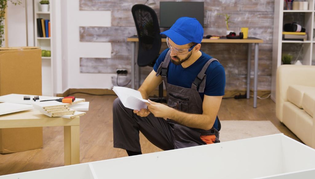 Un homme portant une casquette bleue, des lunettes de protection et une salopette de travail grise est à genoux sur le sol