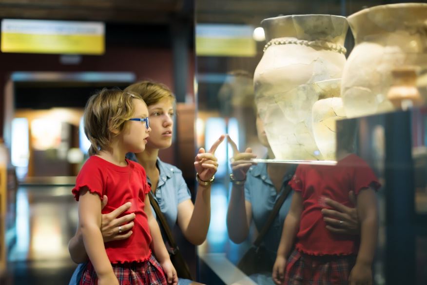 Une femme explique le contenu d'une vitrine à une petite fille portant des lunettes bleues et un t-shirt rouge