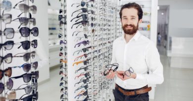 Un opticien barbu en chemise blanche présente plusieurs paires de lunettes devant des présentoirs muraux remplis de montures.