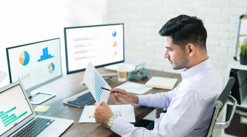 un homme travaille à son bureau devant plusieurs écrans affichant des graphiques financiers et des statistiques