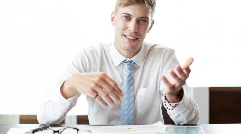 Un jeune consultant souriant en chemise blanche et cravate bleue explique des documents posés sur son bureau
