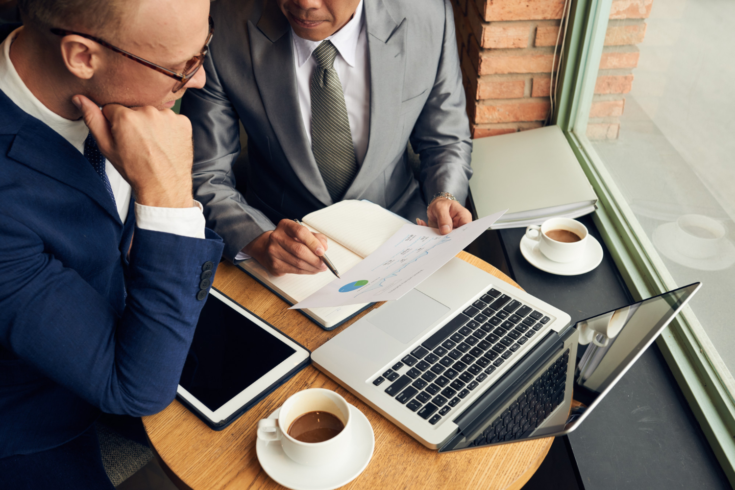 deux hommes d'affaires en costume assis à une table de café, analysant ensemble un document de graphiques financiers à côté d'un ordinateur portable et d'une tablette