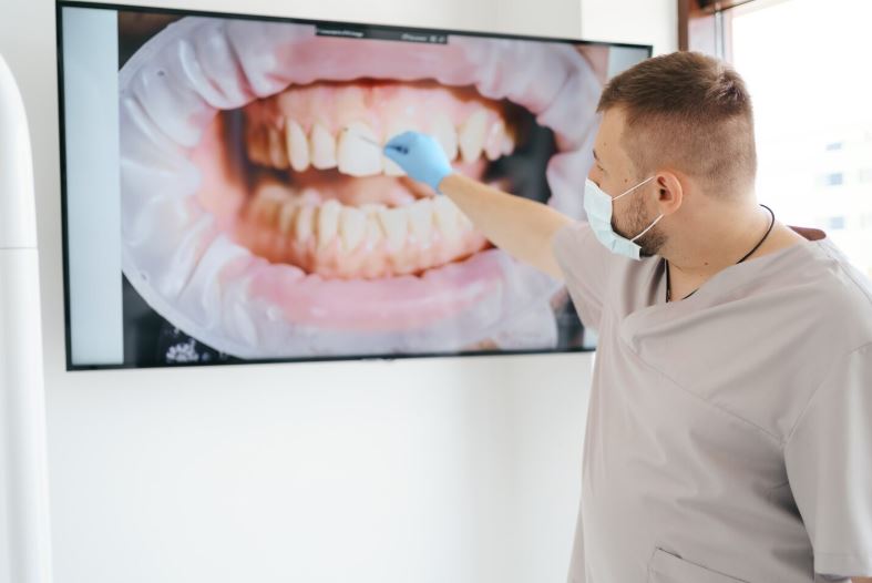 Un dentiste portant un masque chirurgical pointe une radiographie détaillée d'une dentition sur un écran haute définition.