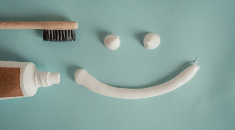 une brosse à dents en bambou et un tube de dentifrice à gauche. Au centre, trois noisettes de dentifrice blanc sous forme d'un visage souriant