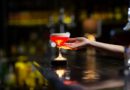 Une main s'apprête à saisir un cocktail rouge vif surmonté d'une mousse blanche, posé sur le comptoir sombre d'un bar rétroéclairé.
