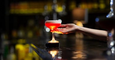 Une main s'apprête à saisir un cocktail rouge vif surmonté d'une mousse blanche, posé sur le comptoir sombre d'un bar rétroéclairé.