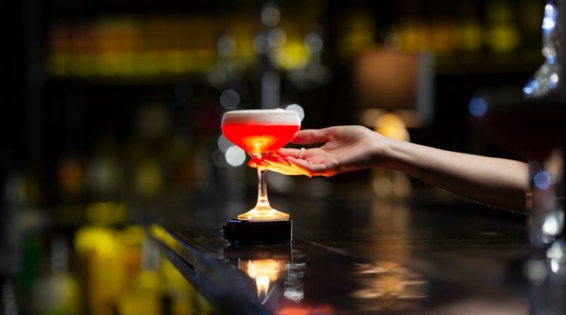 Une main s'apprête à saisir un cocktail rouge vif surmonté d'une mousse blanche, posé sur le comptoir sombre d'un bar rétroéclairé.