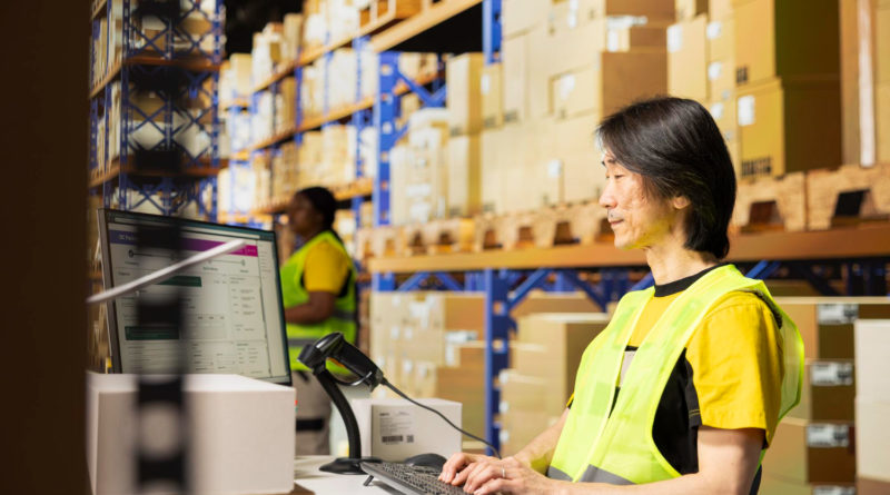Un opérateur logistique asiatique en gilet jaune tape sur son clavier face à son écran de contrôle dans un entrepôt de stockage.