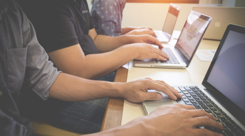les mains de plusieurs personnes assises en ligne, en train de travailler activement sur leurs ordinateurs portables alignés sur une table de bureau