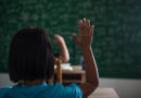 Une élève de dos lève la main en classe face à un tableau vert couvert d'inscriptions floues