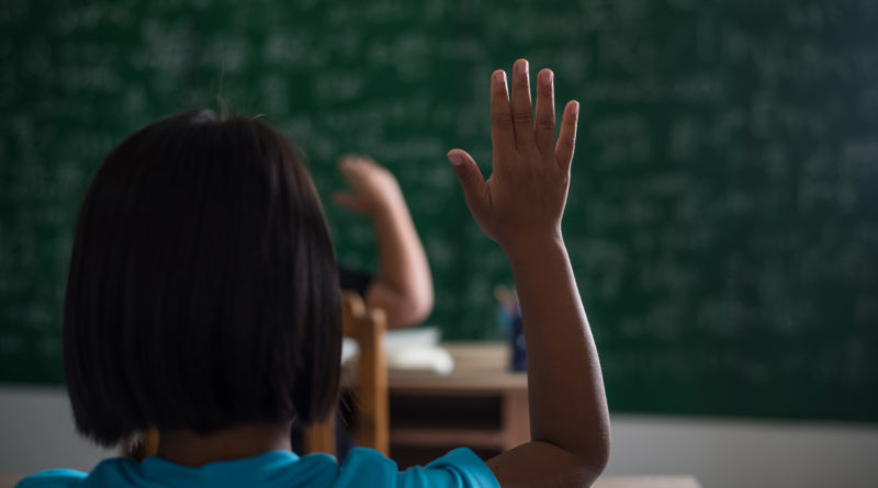 Une élève de dos lève la main en classe face à un tableau vert couvert d'inscriptions floues