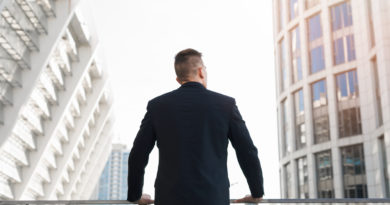 Un homme en costume noir, vu de dos, regarde vers le haut en direction de grands immeubles de bureaux modernes.