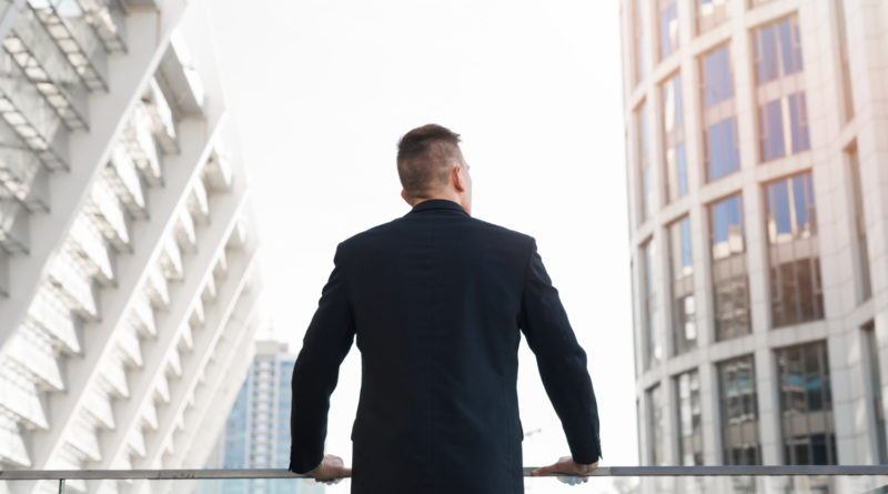 Un homme en costume noir, vu de dos, regarde vers le haut en direction de grands immeubles de bureaux modernes.