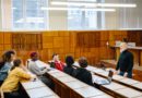 un enseignant senior à la barbe blanche debout face à un groupe d'étudiants multiculturels attentifs lors d'un cours dispensé dans un amphithéâtre universitaire.