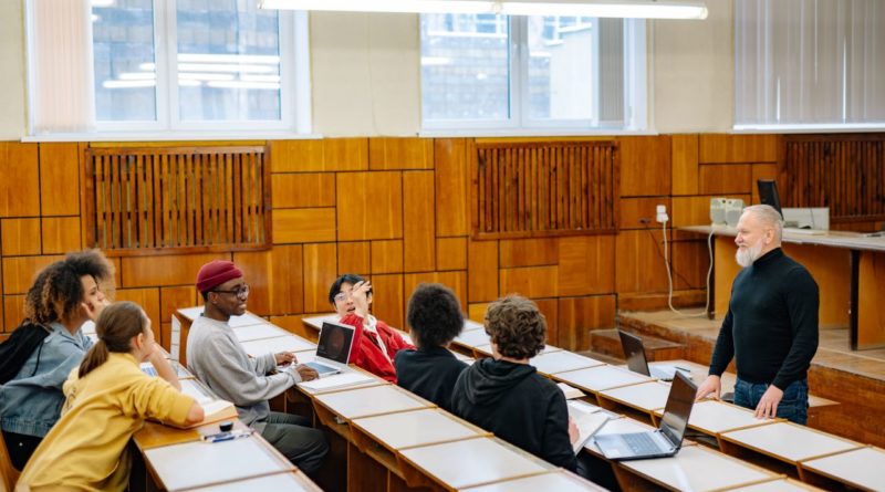 un enseignant senior à la barbe blanche debout face à un groupe d'étudiants multiculturels attentifs lors d'un cours dispensé dans un amphithéâtre universitaire.