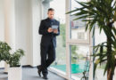 Portrait d'un dirigeant d'entreprise en costume analysant des données sur sa tablette tactile, debout près d'une fenêtre de bureau avec des plantes vertes en arrière-plan.