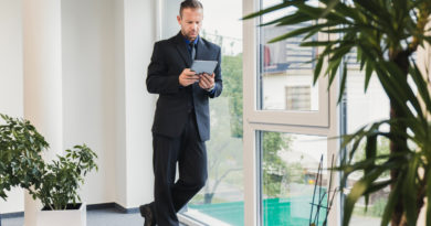 Portrait d'un dirigeant d'entreprise en costume analysant des données sur sa tablette tactile, debout près d'une fenêtre de bureau avec des plantes vertes en arrière-plan.