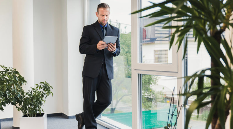 Portrait d'un dirigeant d'entreprise en costume analysant des données sur sa tablette tactile, debout près d'une fenêtre de bureau avec des plantes vertes en arrière-plan.