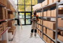 un employé de logistique debout au milieu des étagères remplies de cartons dans un entrepôt, une tablette numérique à la main pour gérer les stocks