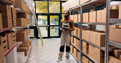 un employé de logistique debout au milieu des étagères remplies de cartons dans un entrepôt, une tablette numérique à la main pour gérer les stocks