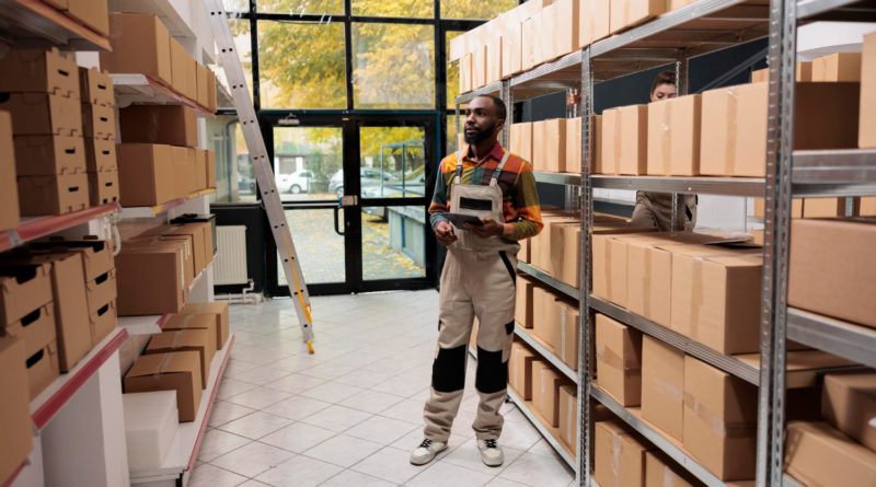 un employé de logistique debout au milieu des étagères remplies de cartons dans un entrepôt, une tablette numérique à la main pour gérer les stocks